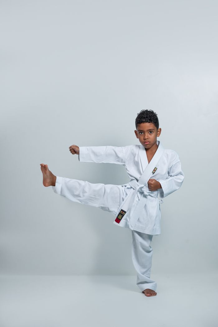 A young boy demonstrates a karate kick in a studio setting, wearing a white kimono.
